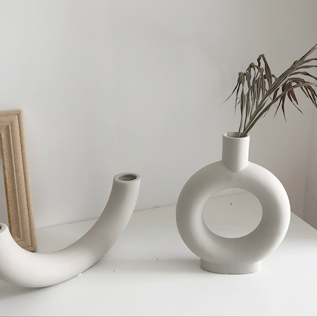 Half pipe vase resting beside a round doughnut vase holding dried palm, against a pale wall