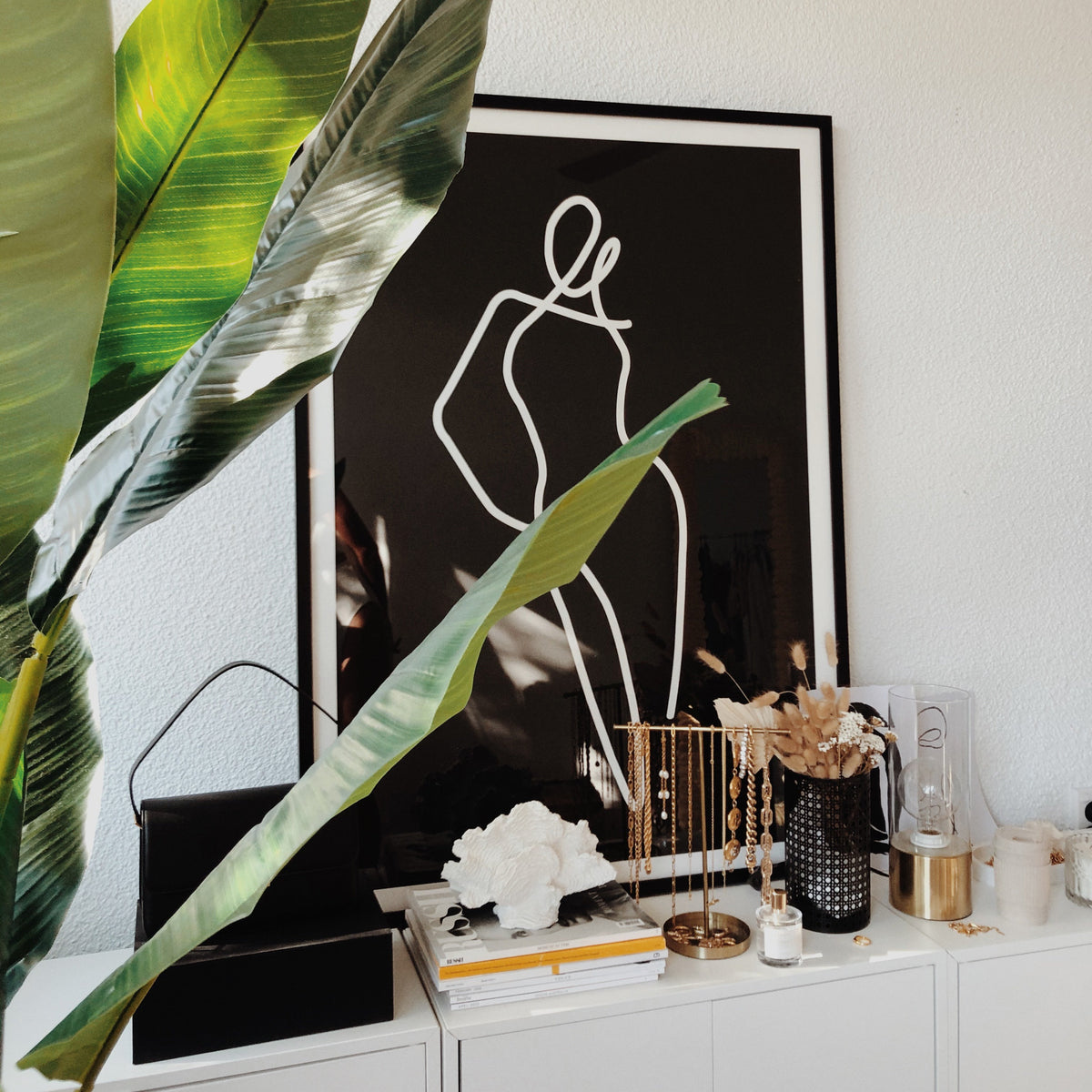 Homebodii Black print in a black frame on a white console beside a banana leaf and jewellery