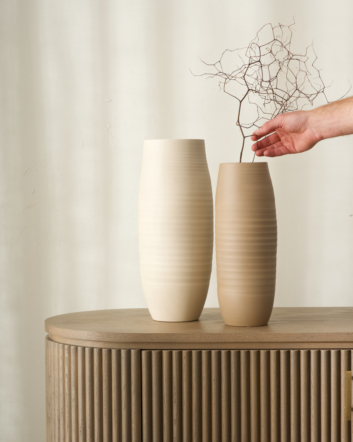 Large Beige and Small Mud vases together, a hand placing a dried branch into the sand coloured vase