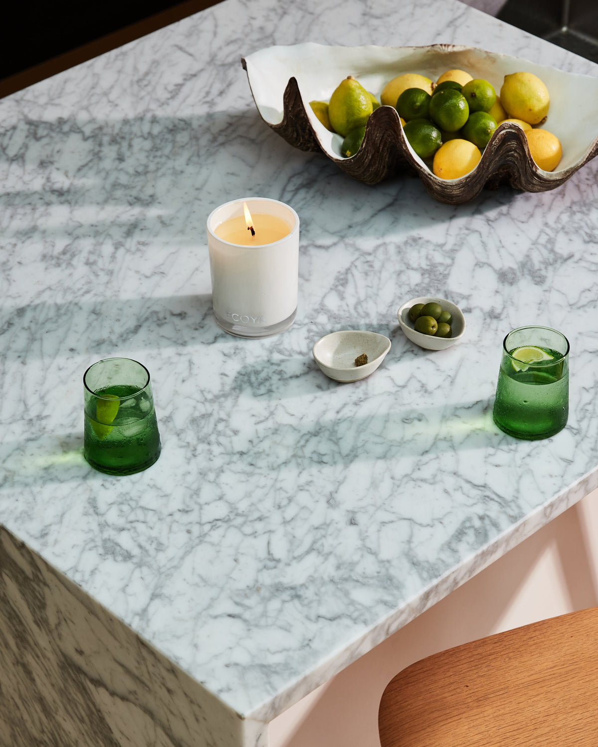 A lit Ecoya Madison candle on a marble benchtop with green glasses and a shell bowl of limes and lemons