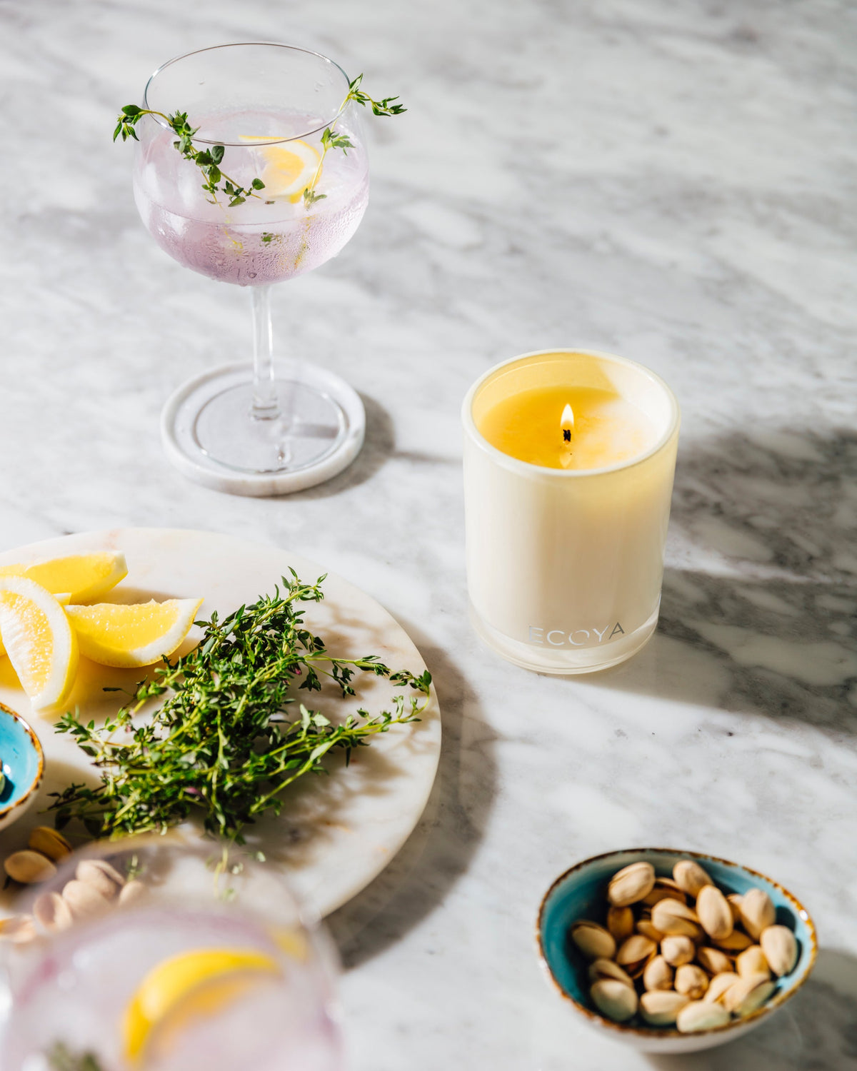 Lit Ecoya Madison candle on a marble bench with sliced citrus, herbs and a bowl of nuts