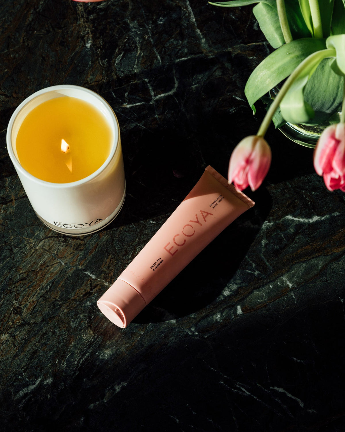 Peach Ecoya hand cream beside a lit candle and pink tulips on dark marble