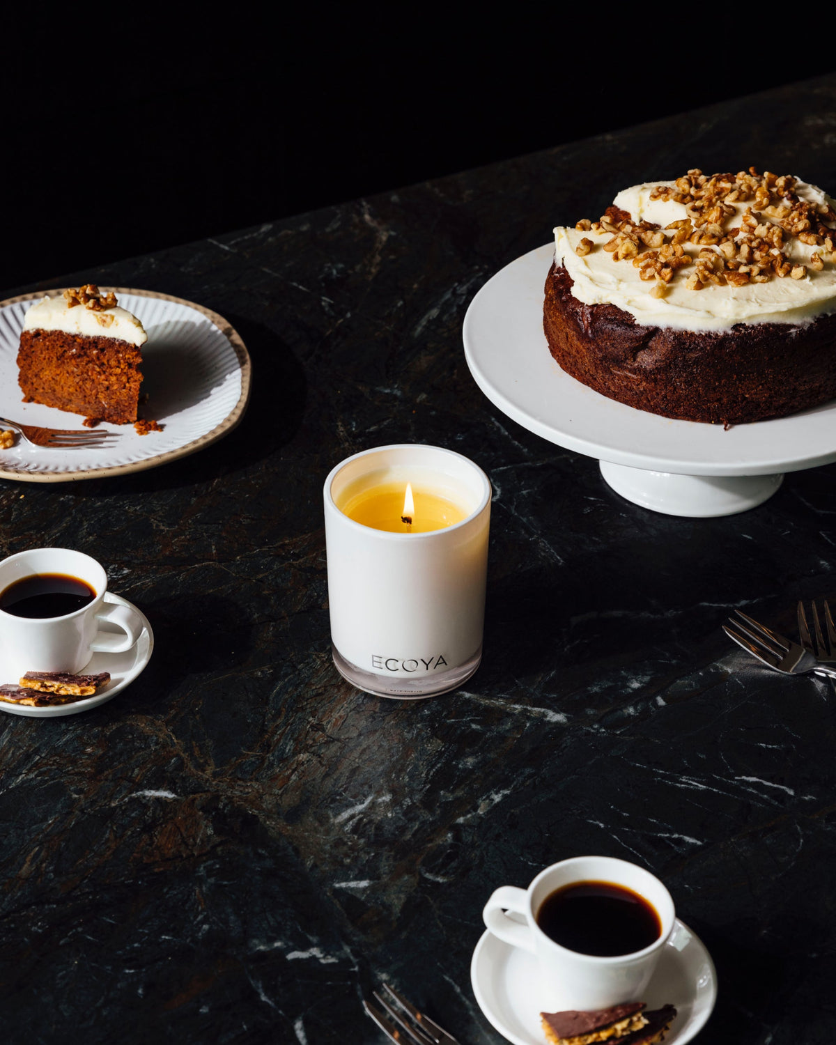 A lit Ecoya Madison candle on a dark marble table with a frosted layer cake, espresso cups and a slice on a plate