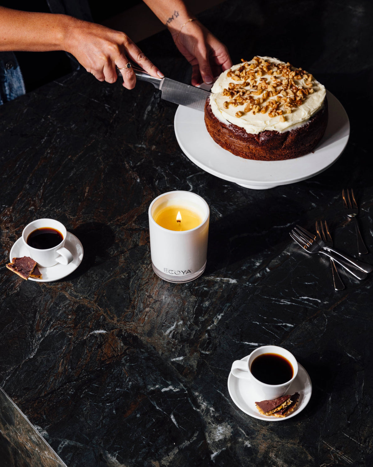 A lit Ecoya Madison candle on a dark marble table while someone slices a frosted cake, with espresso cups alongside