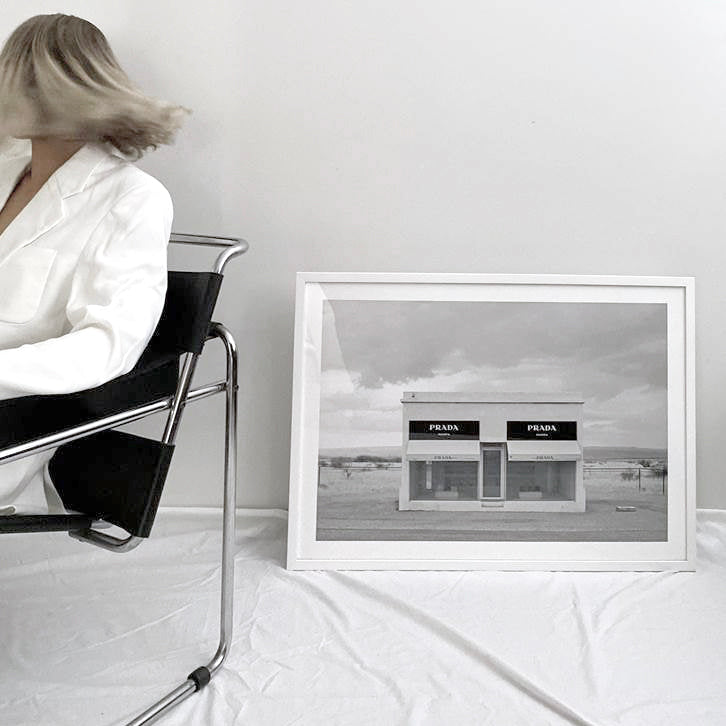 Marfa print in a white frame leaning on the floor beside a woman on a black chair