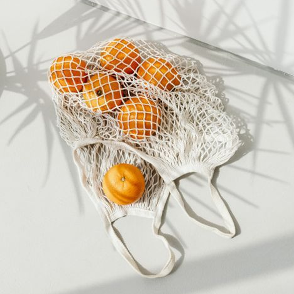 Cream string bag full of mandarins on a white surface under the shadow of a palm frond
