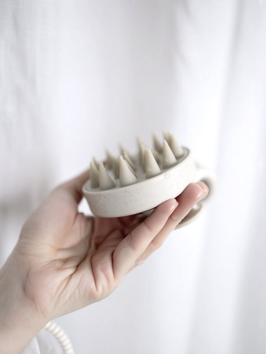 A hand holding the speckled cream scalp massager against a white linen curtain