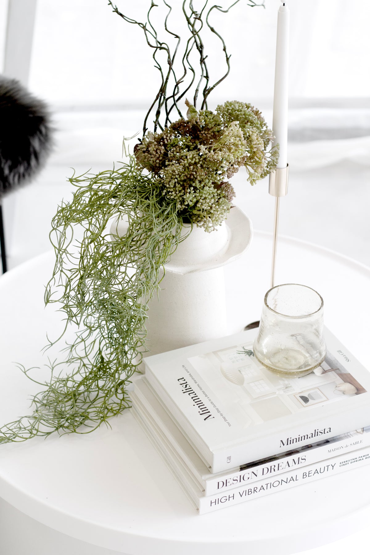 Minimalista by Shira Gill book styled on white table with sculptural vase, greenery and candle