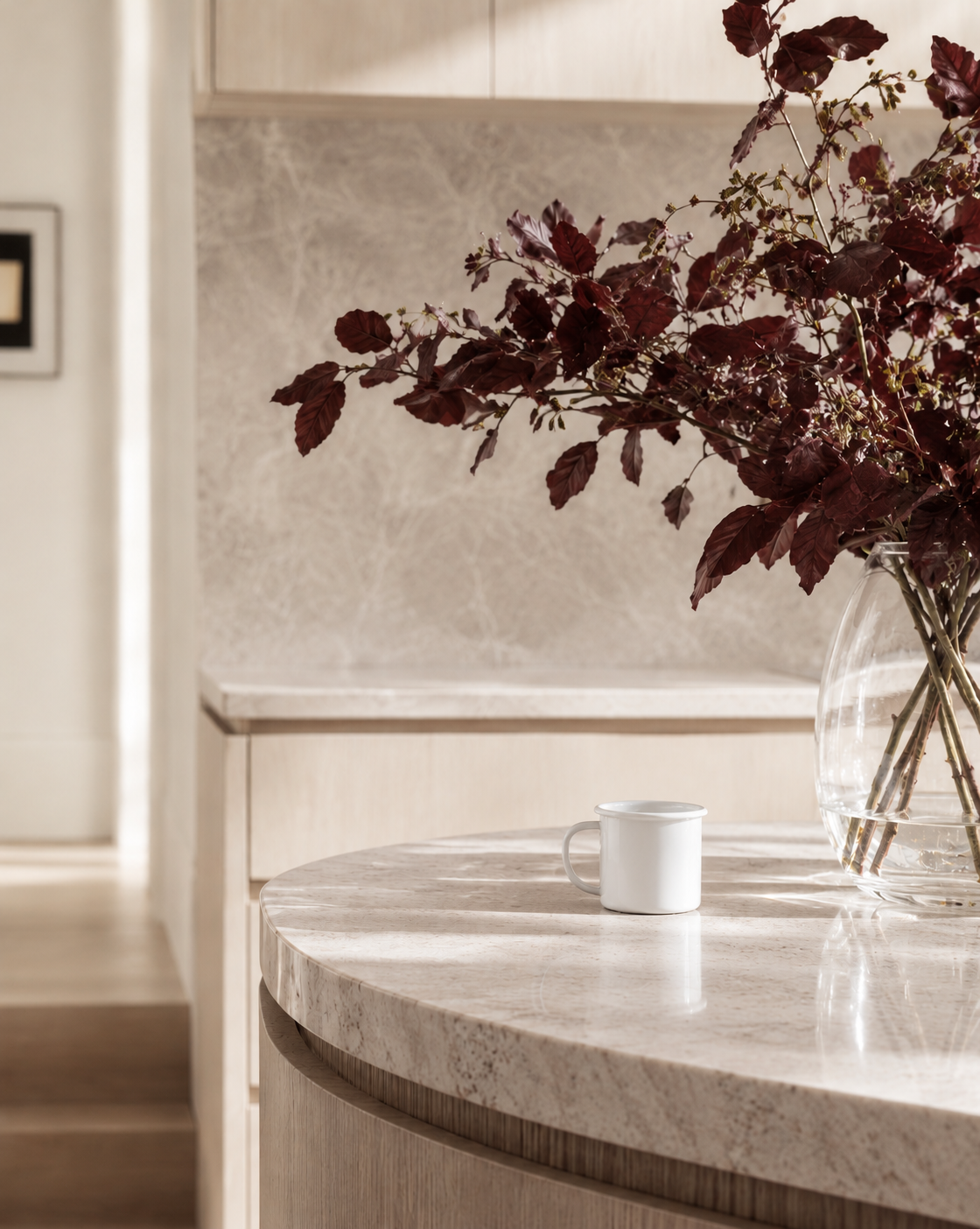 Small white enamel mug on a curved stone island beside a vase of copper beech leaves
