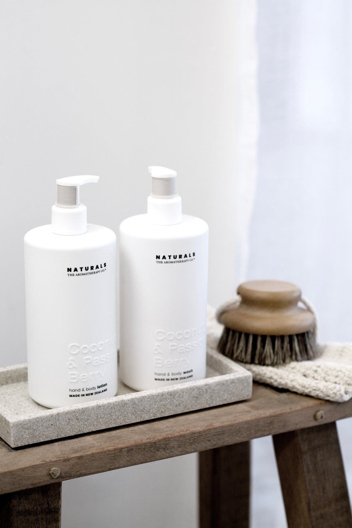 Two Naturals pump bottles on a stone tray on a rustic timber stool with a bath brush