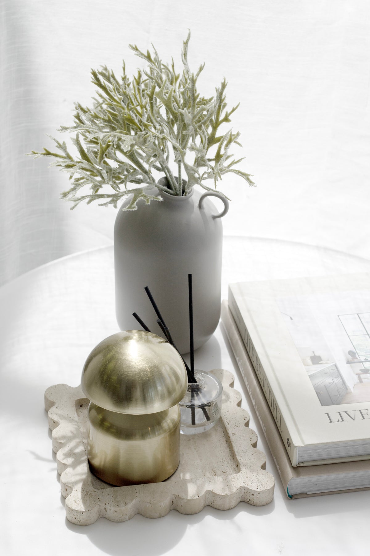 Grey Flugen vase with soft green foliage beside a gold candle and books on a white table