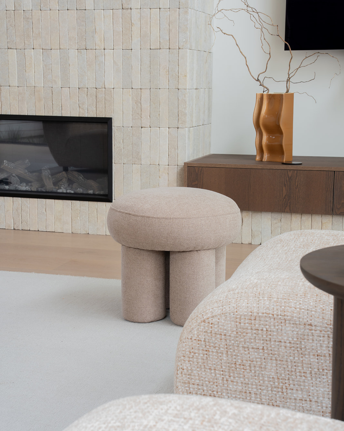 Wave-folded vase with twisting branches on a floating timber shelf beside a fireplace and bouclé stool
