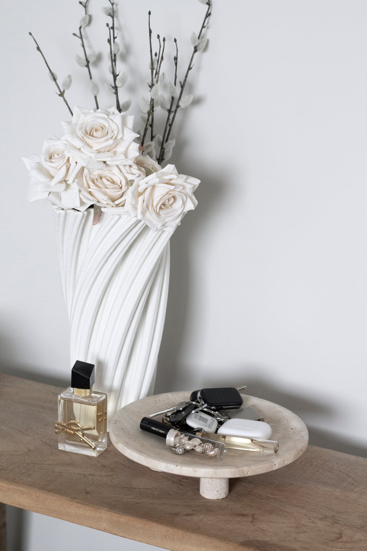 Nome stand holding perfume and keys on a timber console beside a white swirl vase of blossom branches