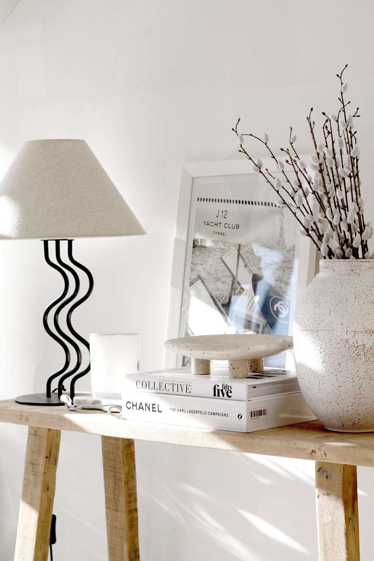Nome stand on a timber side table under a wavy lamp with books, a framed print and dried stems