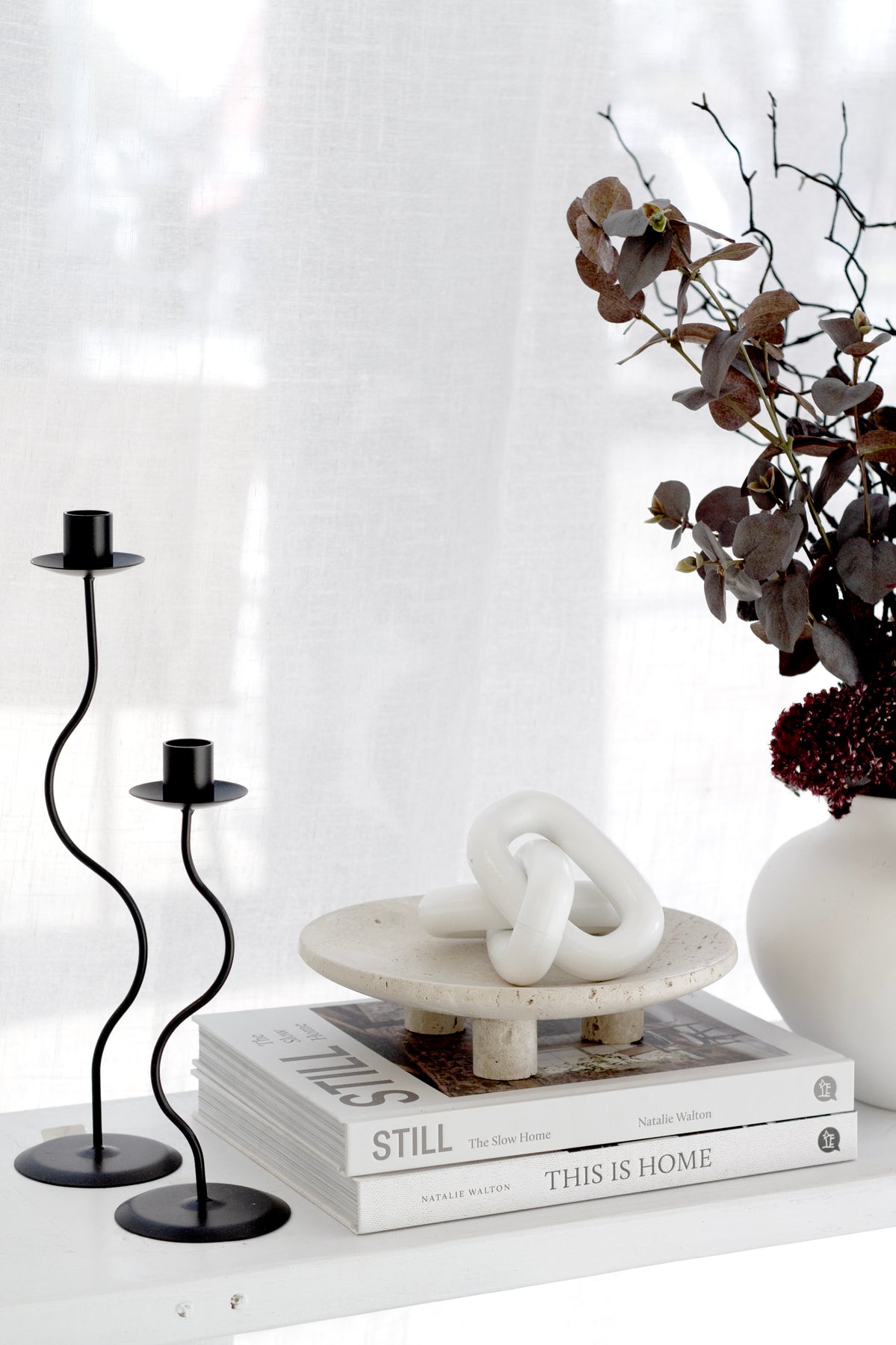 Nome stand holding a white knot sculpture on books beside wavy black candlesticks and dark foliage