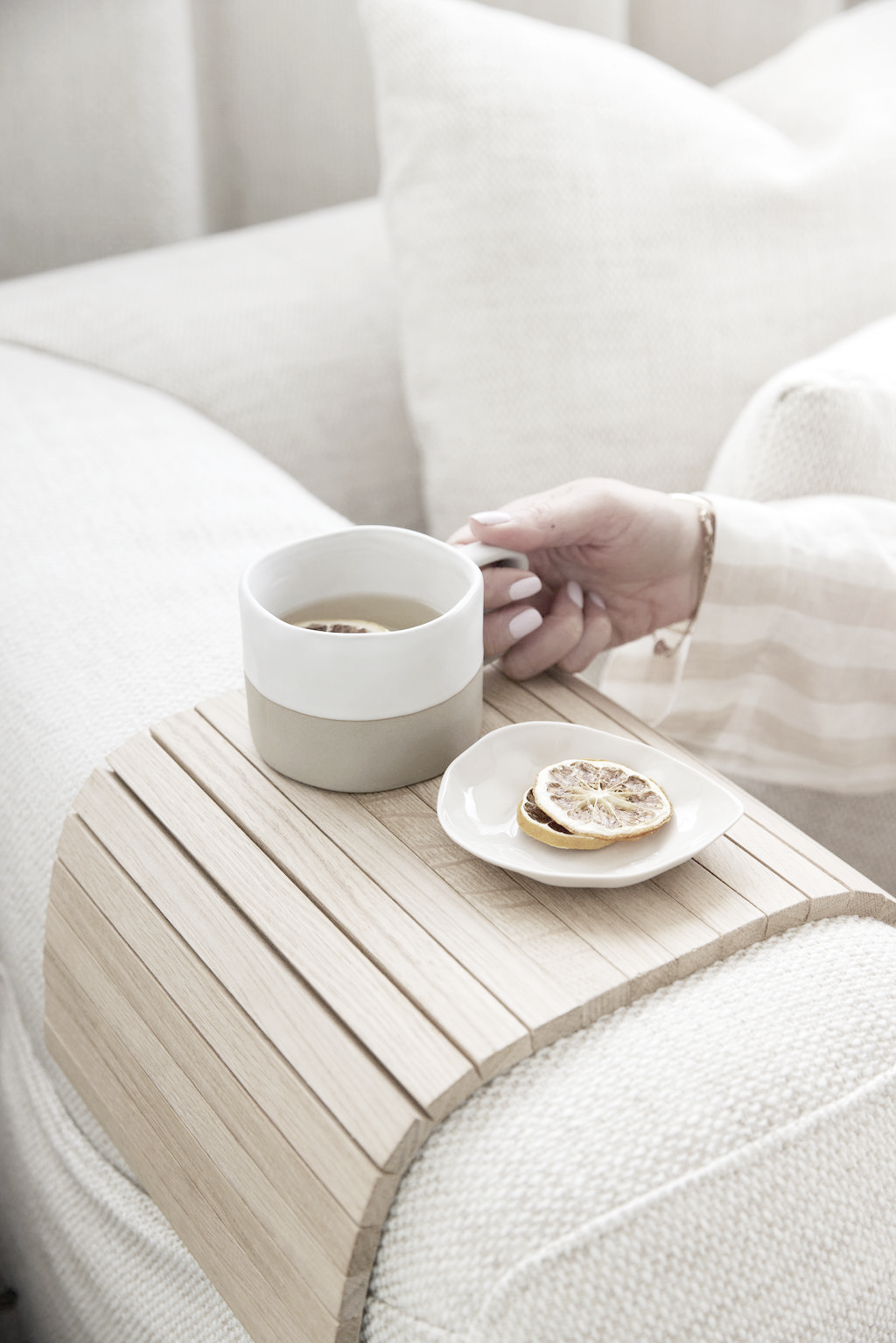 Natural oak armchair tray holding a ceramic mug and a small plate of dried lemon slices on a neutral sofa