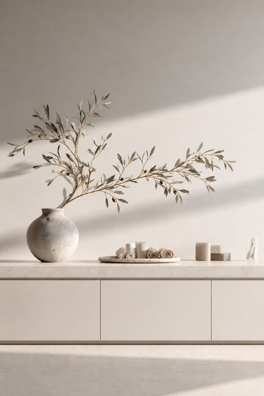 Faux olive branch with small olives arching from a round stone vase on a pale sideboard