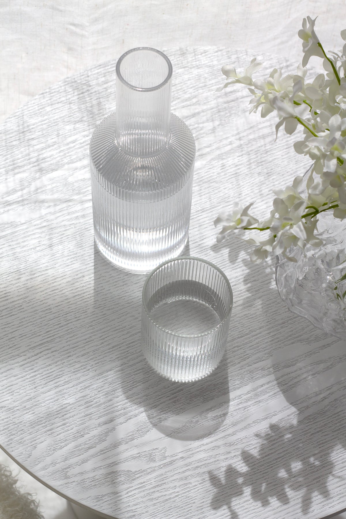 Papier HQ Amalfi fluted glass water decanter and tumbler on white oak table with white orchid blooms and shadow play