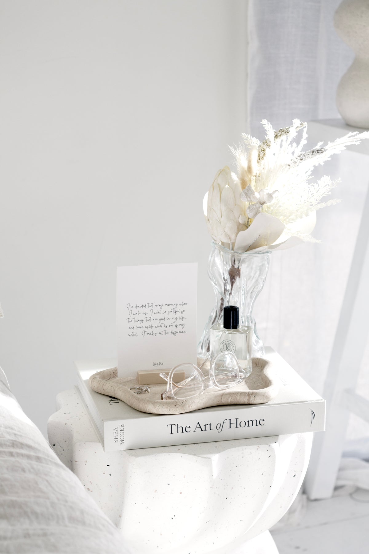 Papier HQ Travertine Curved Tray styled on a white side table with a wavy glass vase of dried white florals, gratitude print card, and The Art of Home book by Shea McGee, airy white bedroom setting.