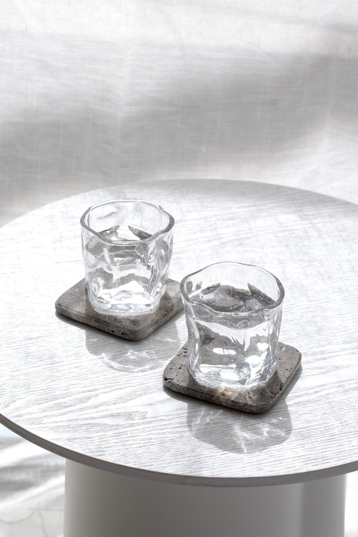 Set of two clear wavy textured tumblers on stone coasters, styled on a round white-washed side table in bright natural light