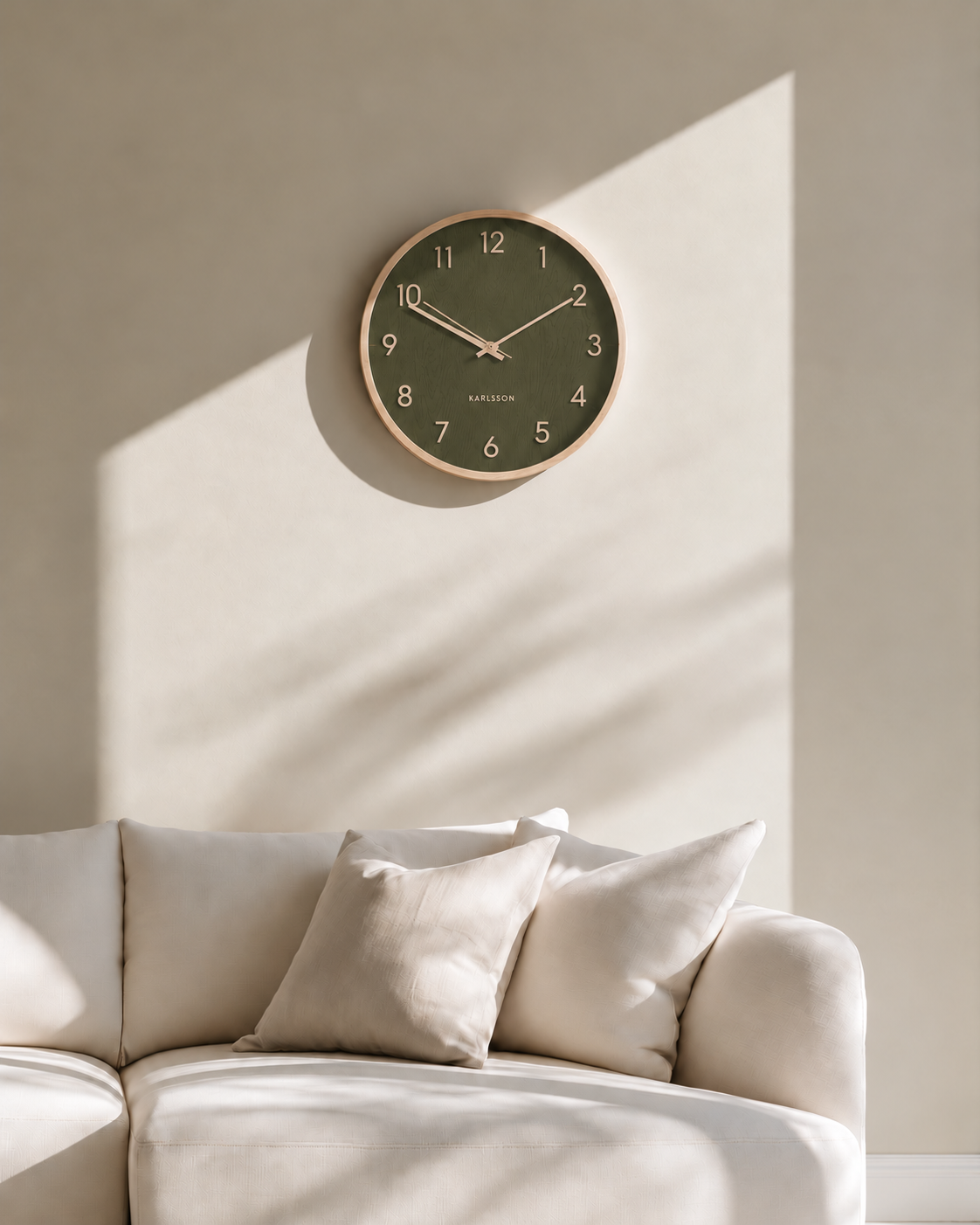 Pure Wood Grain Wall Clock in moss green with light wood rim above a cream sofa in afternoon sun