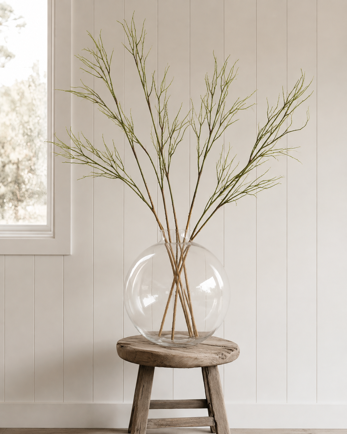 Faux saltmarsh stems in a round clear glass vase on a rustic stool against white panelling