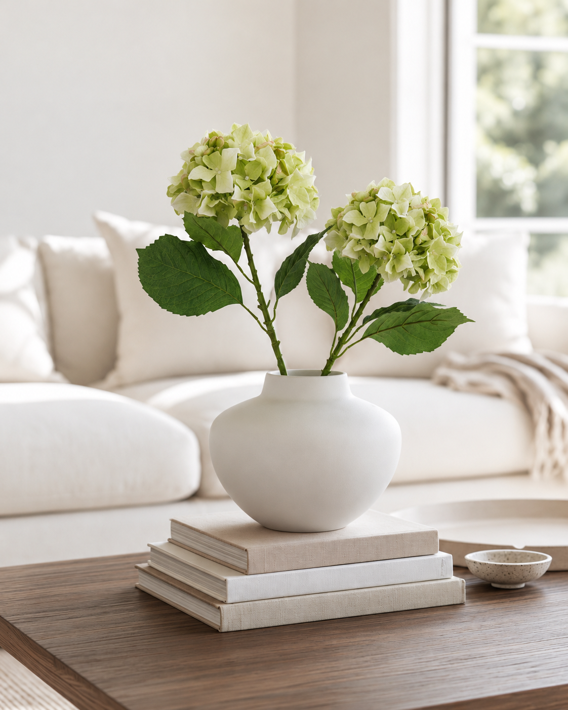 Two green Sublime Hydrangea stems in a matte white vase on stacked books in a bright cream living room