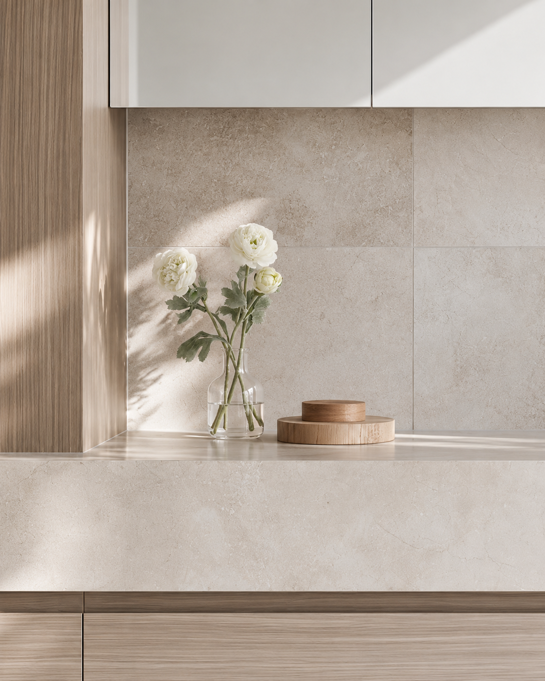 White faux ranunculus stems in a small clear glass vase on a stone bathroom vanity beside a timber dish