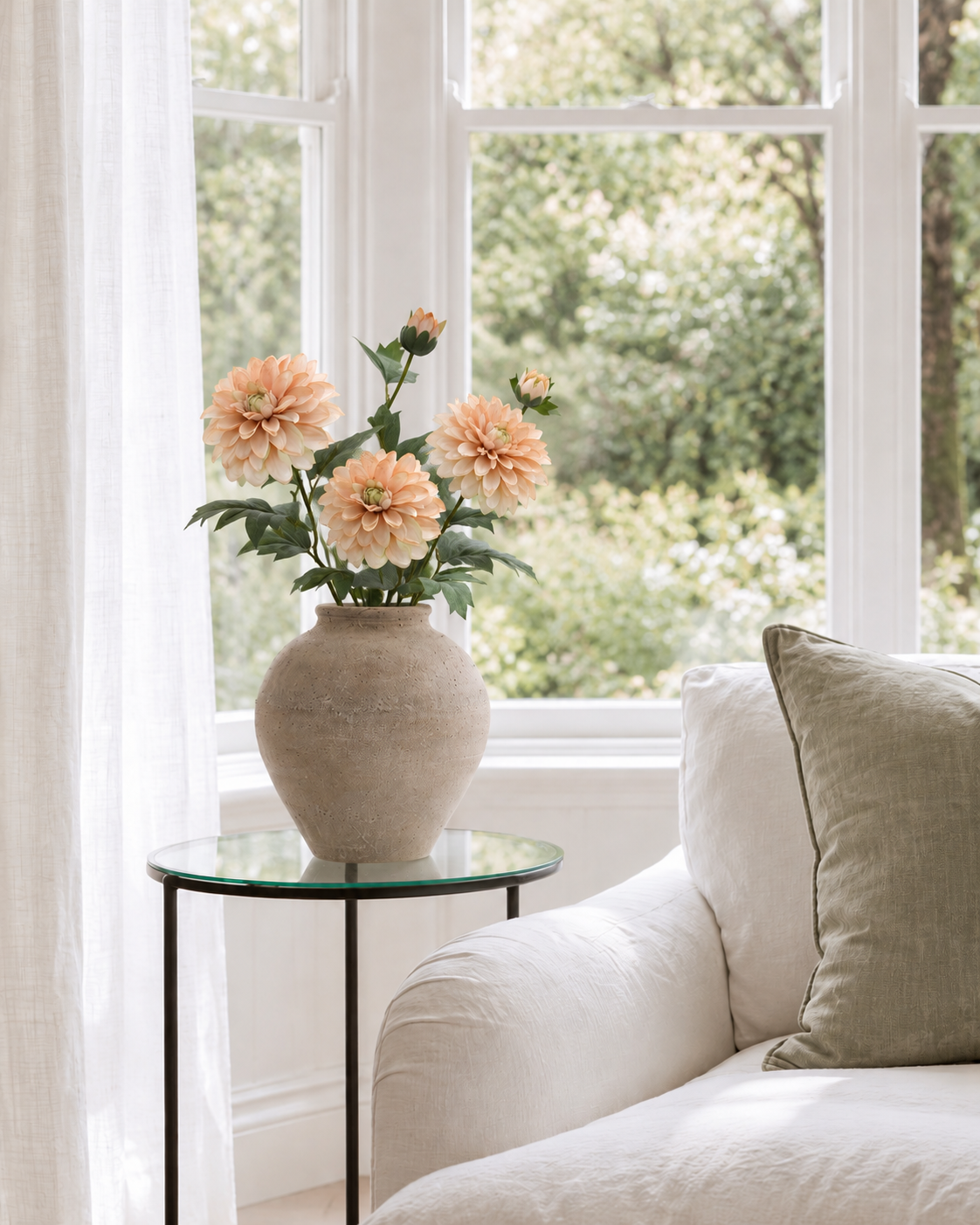 Heritage Dahlia stems in apricot glow in a stone vase on a glass side table by a bay window and linen armchair