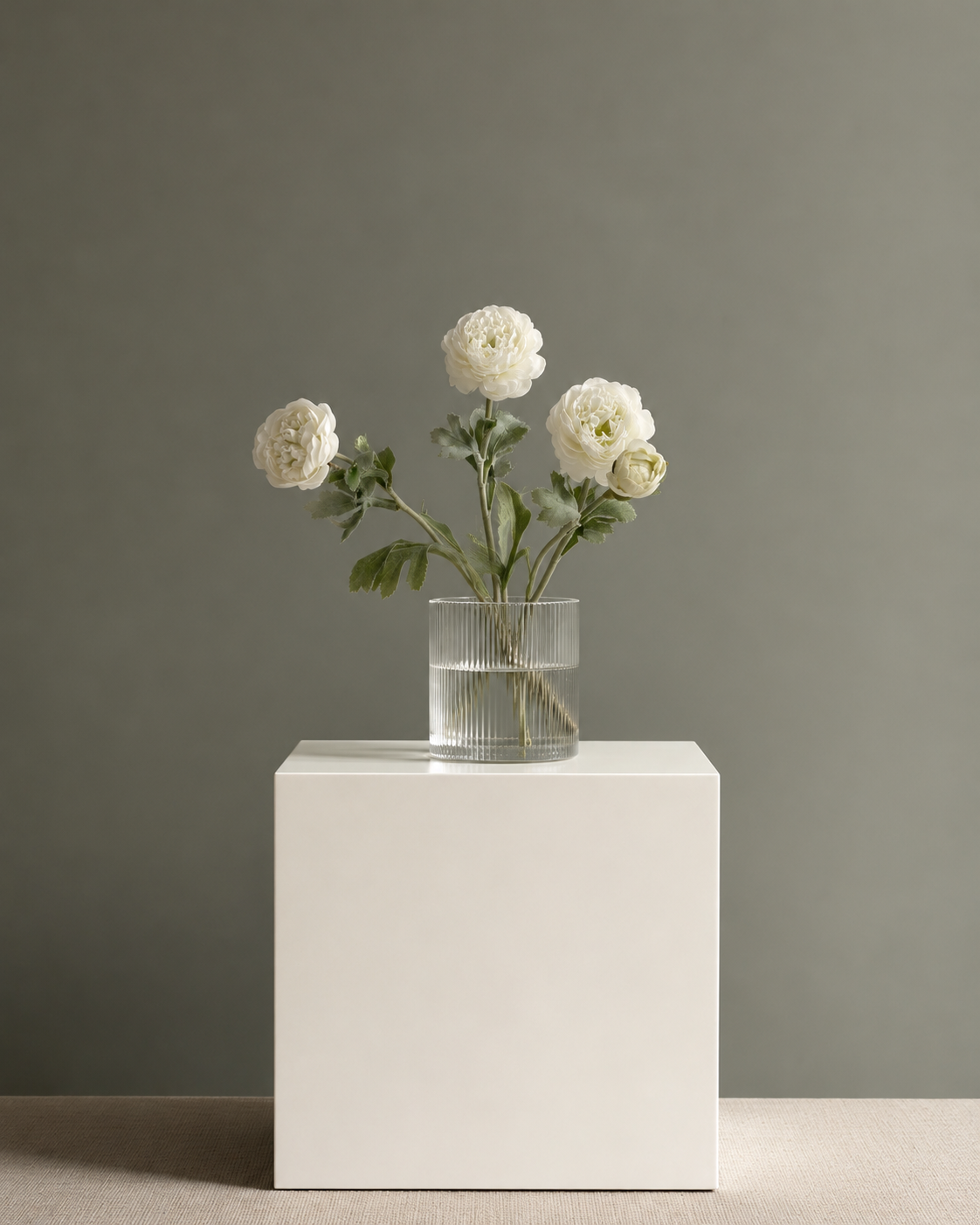 White faux ranunculus stems in a ribbed glass vase on a white plinth against a sage wall