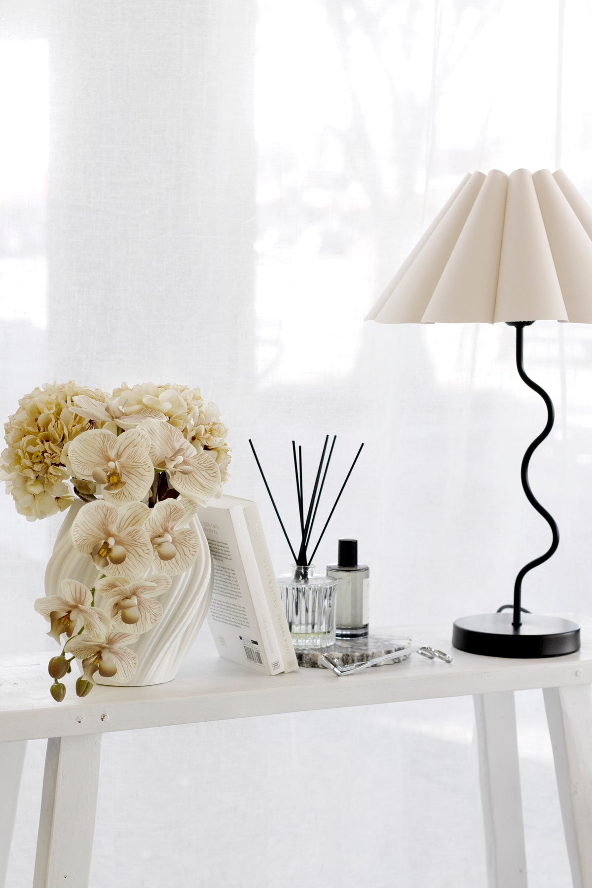 Console styled with orchids in a sculptural vase, a reed diffuser, scent bottles and a lamp