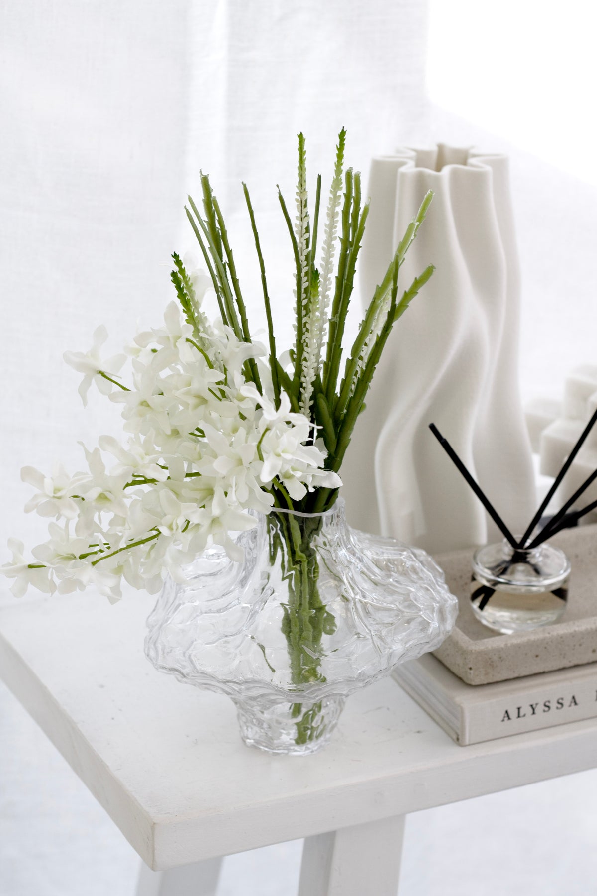 Slender faux rhipsalis stem with white floral detail styled in a sculptural clear glass vase on a white table with soft morning light.