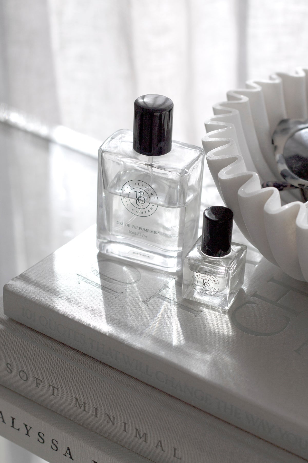 Square perfume oil bottles on neutral books by a sunlit window beside a sculptural flower