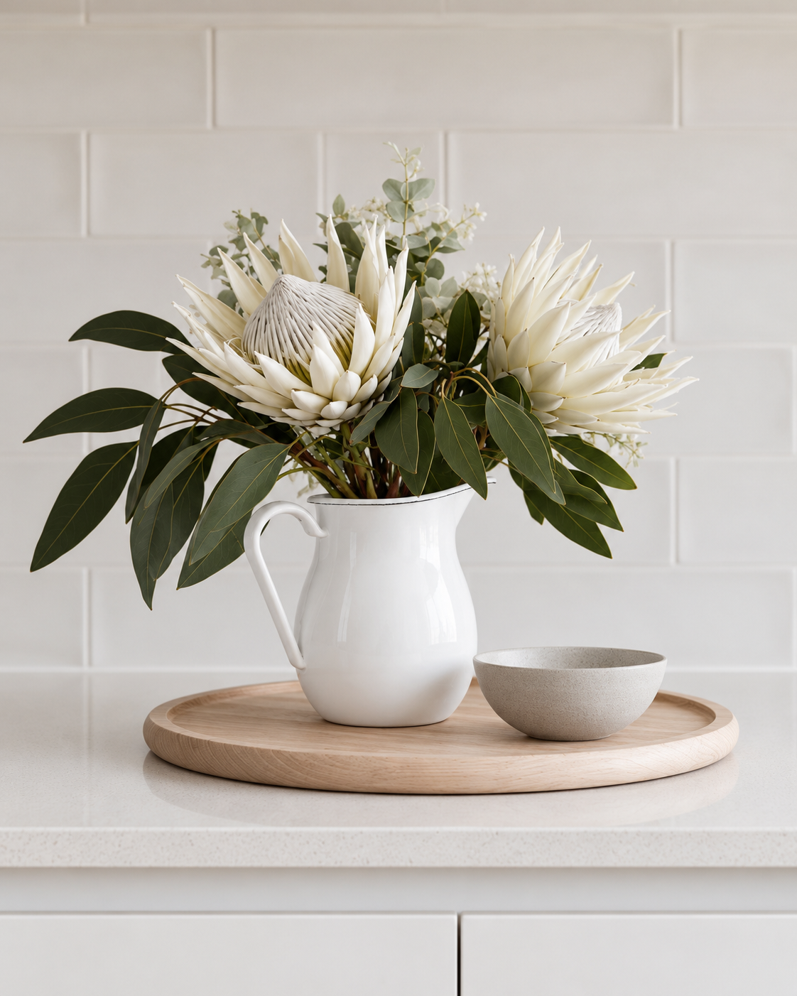 White enamel water jug holding white proteas and eucalyptus on a timber tray against a subway tile splashback