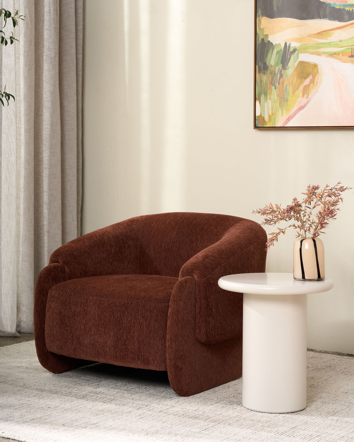 Pale pedestal side table holding a striped vase of dried stems beside a rust-brown armchair