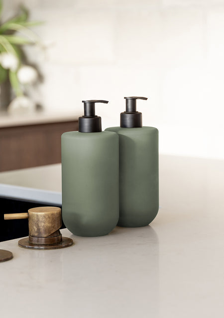 Two sage green Amber & Freesia pump bottles on a white stone benchtop beside a brass tap