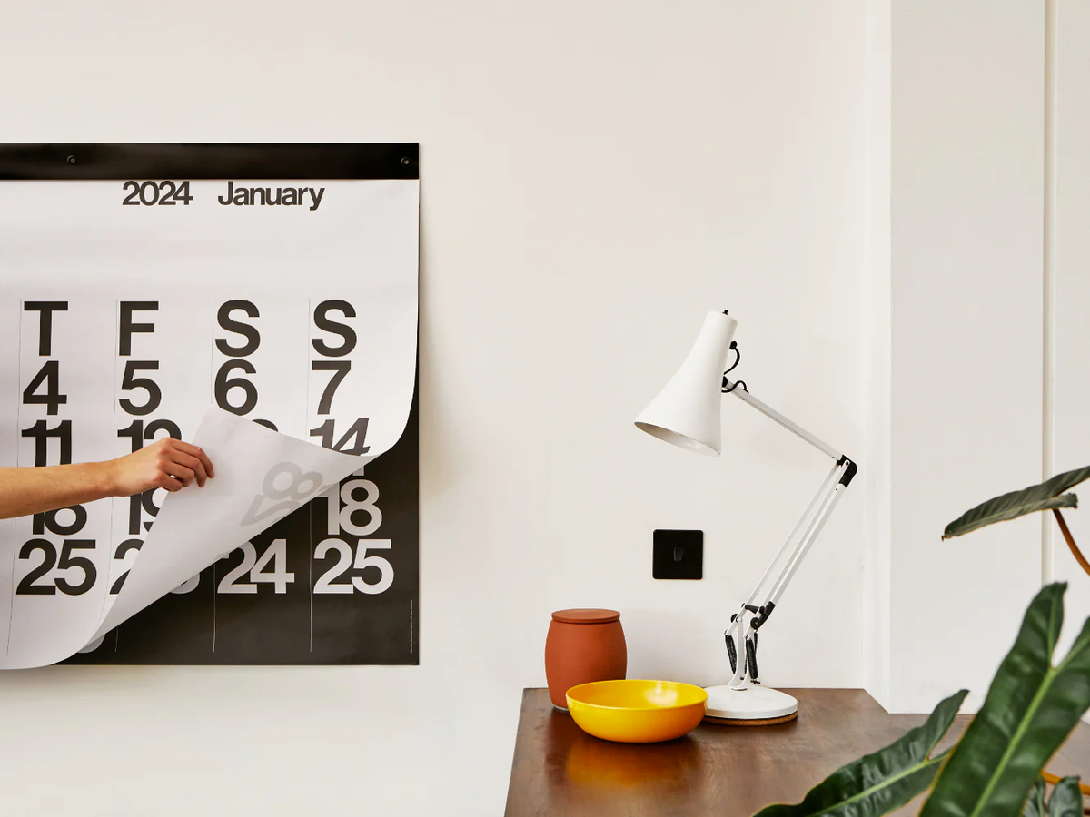 Hand tearing off a monthly page of the Stendig Calendar by Massimo Vignelli above a desk with a white anglepoise lamp