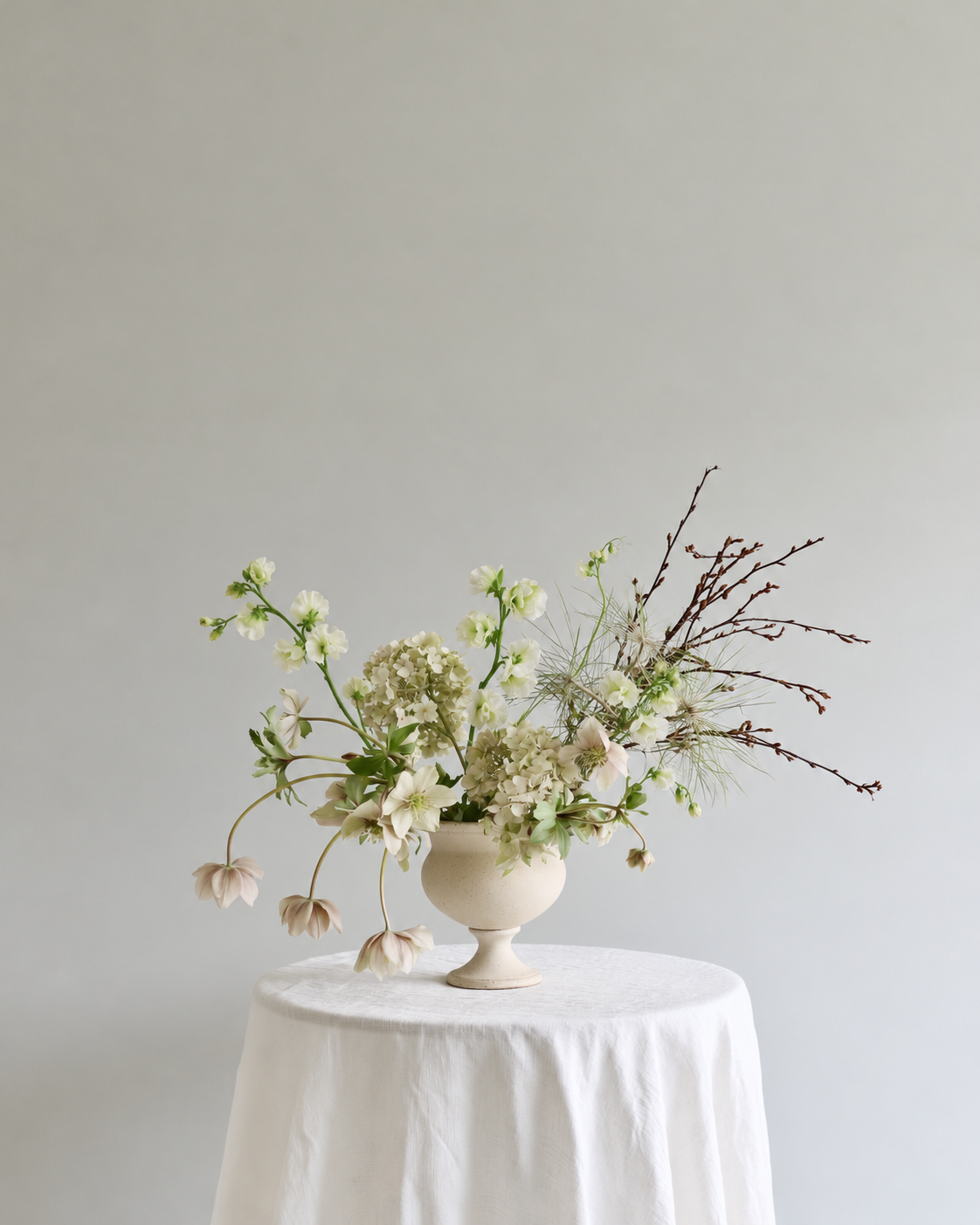 Cream sweet pea stems woven into a loose garden arrangement in a white footed vase on a draped table