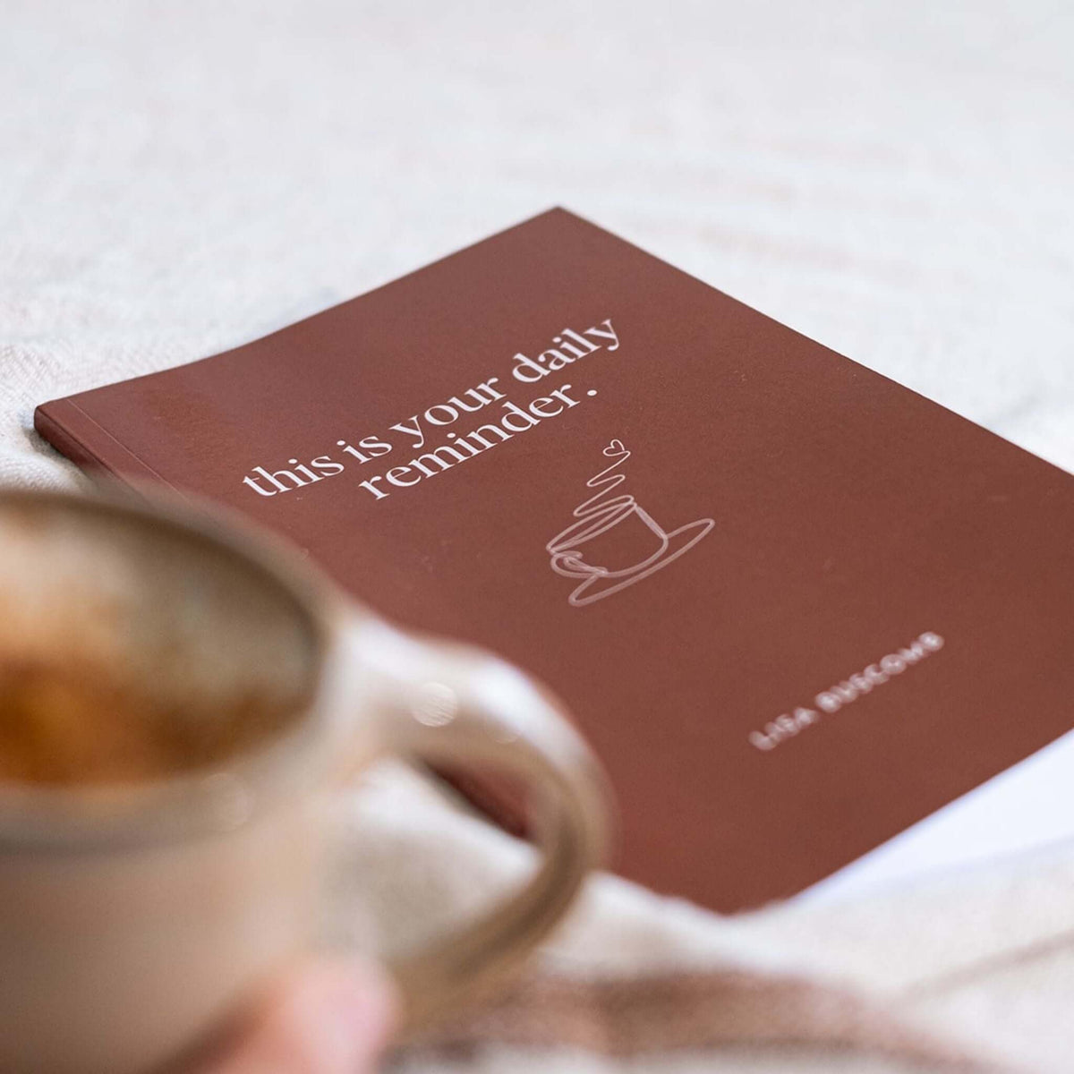 This Is Your Daily Reminder book by Lisa Buscomb with a terracotta cover, behind a ceramic coffee mug