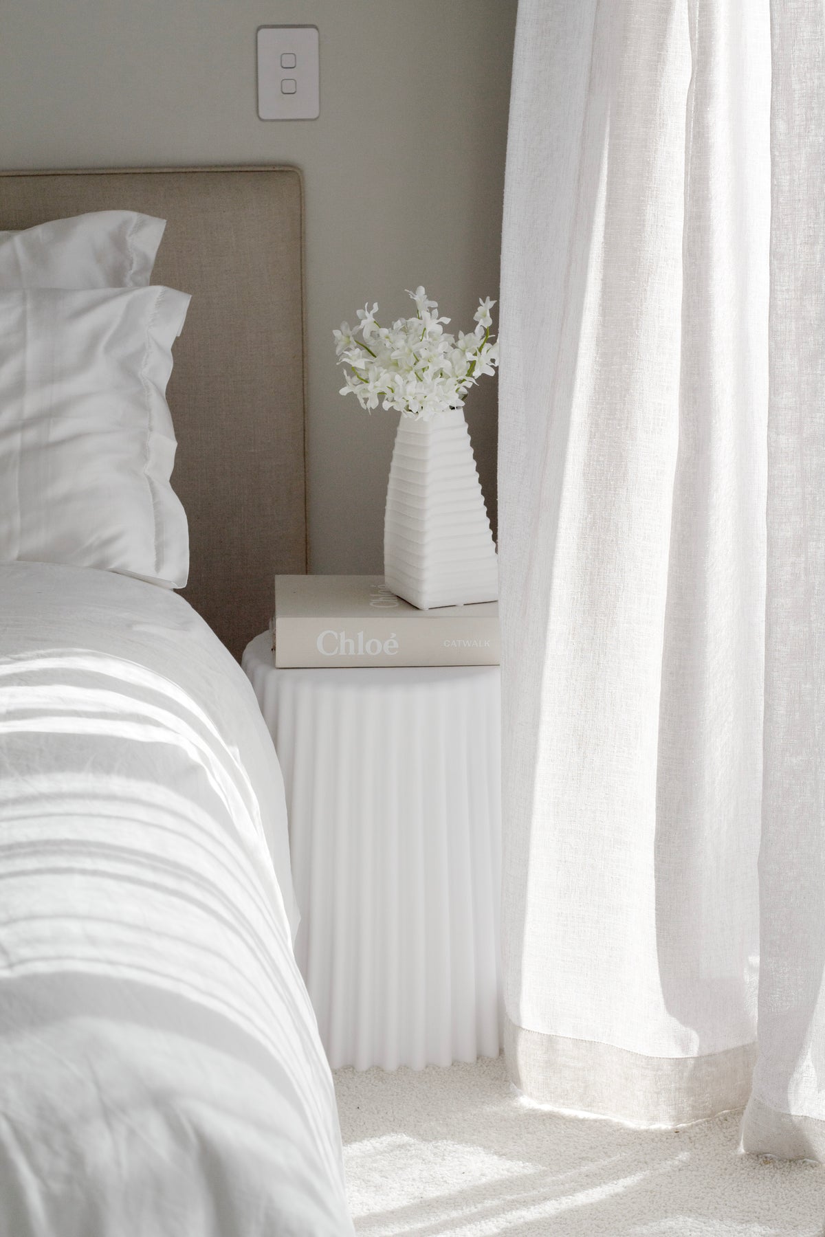 White ribbed polyurethane stool styled as bedside table beside neutral bed and sheer curtains