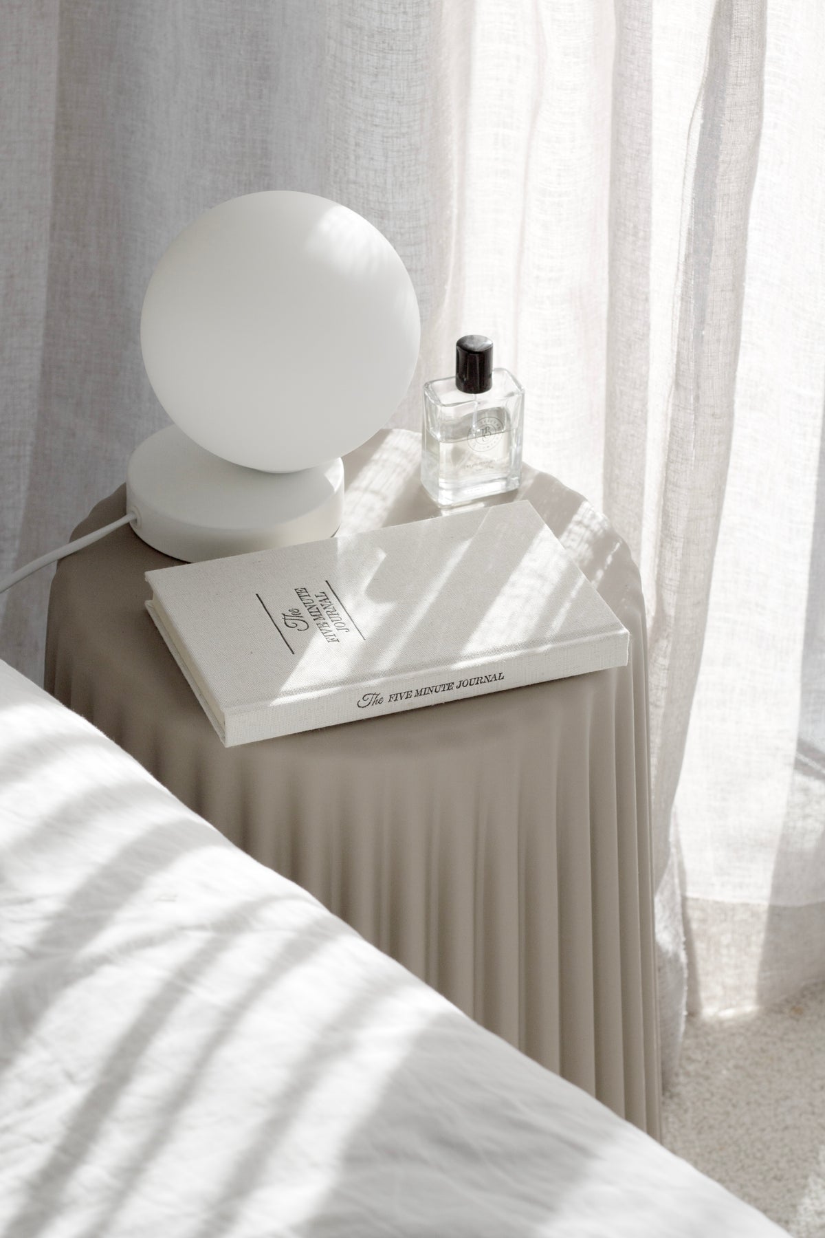 White ribbed stool as a bedside table with a globe lamp and a small bottle, sun striping the curtain