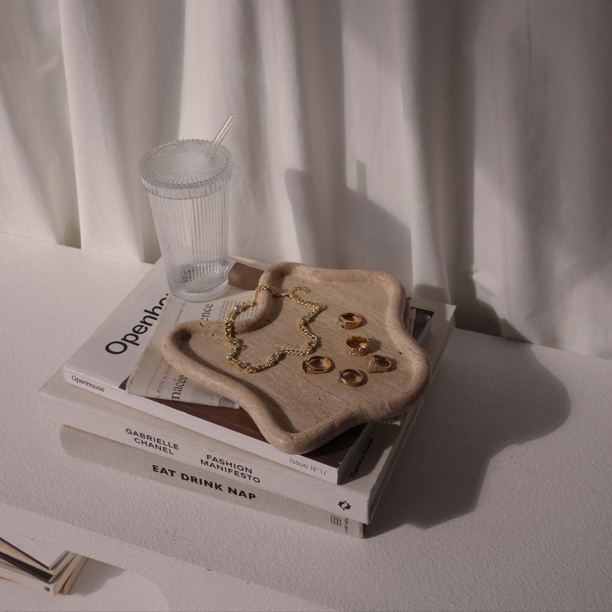 Travertine curved tray holding gold jewellery on a stack of books by a sheer curtain