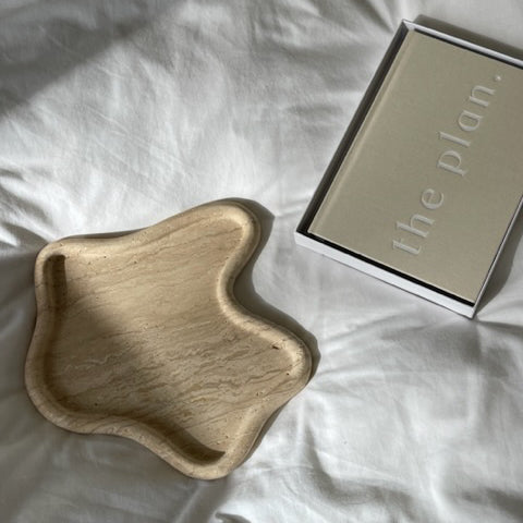 Travertine curved tray on white bed linen beside a notebook