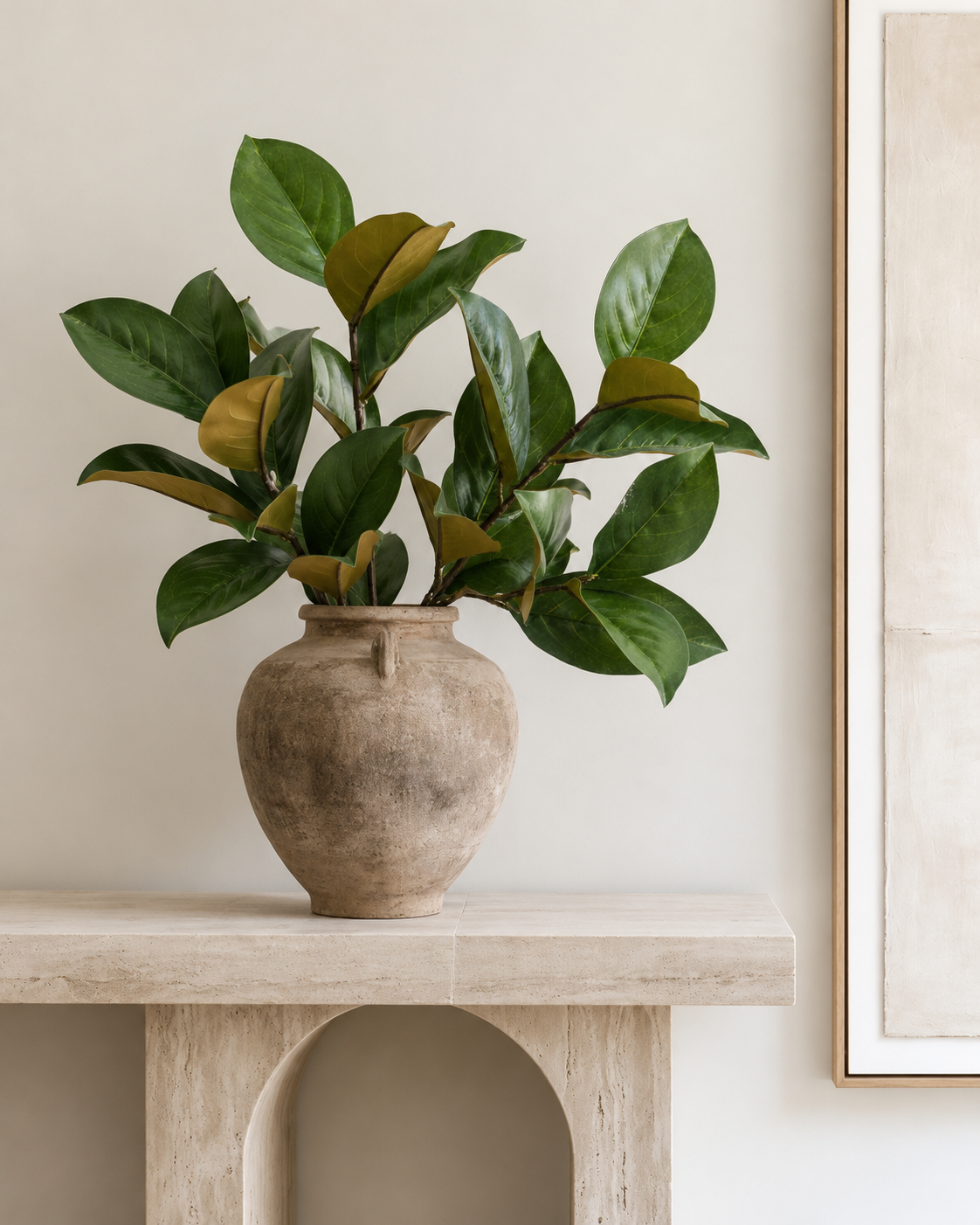 Faux magnolia leaf sprays in a rustic clay urn on a travertine console beside framed art