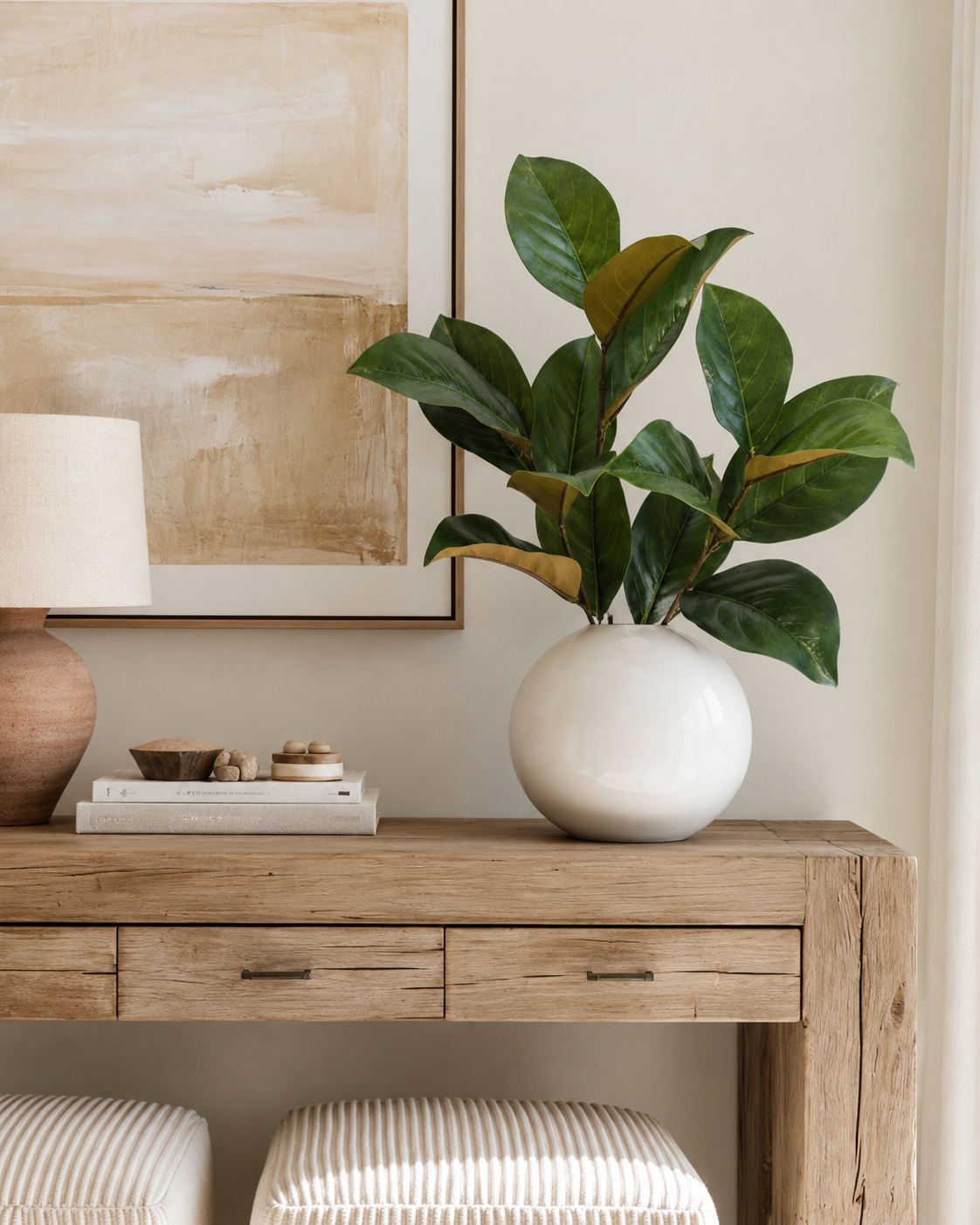 Faux magnolia leaf sprays in a round white vase on a weathered oak console with a lamp and abstract art