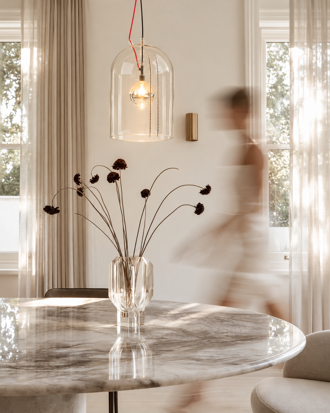 Milk Jar clear glass pendant glowing above a round marble dining table with dark flower stems in a glass vase
