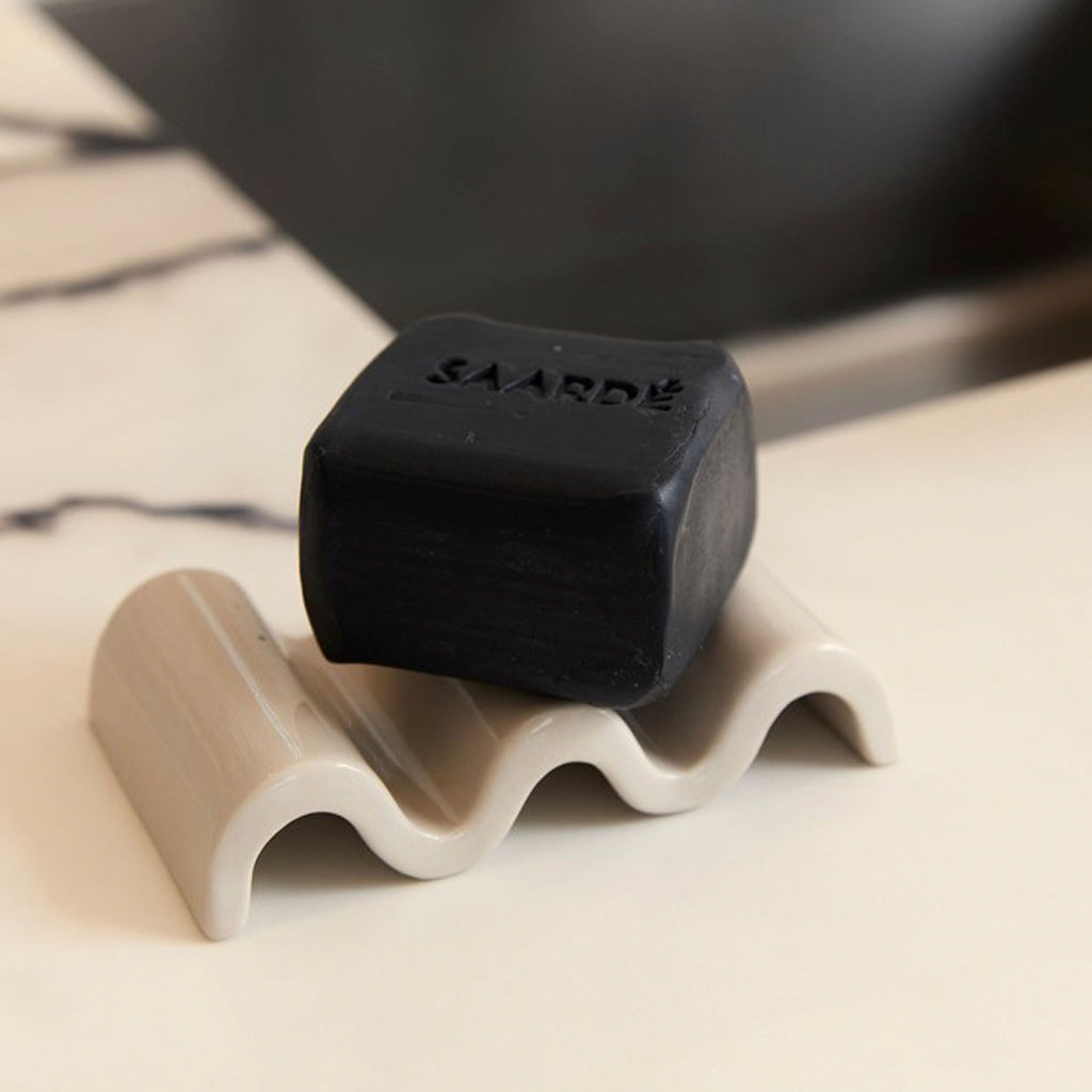 Wavy ceramic ripple dish in a soft taupe glaze used as a soap holder on a marble bathroom bench with a black bar of soap.