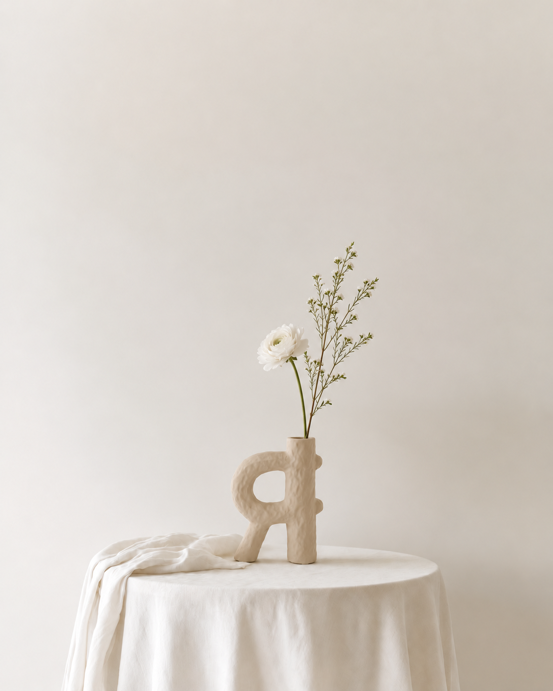 Weir Textured Vase in sand with a single white ranunculus and waxflower on a linen-draped side table