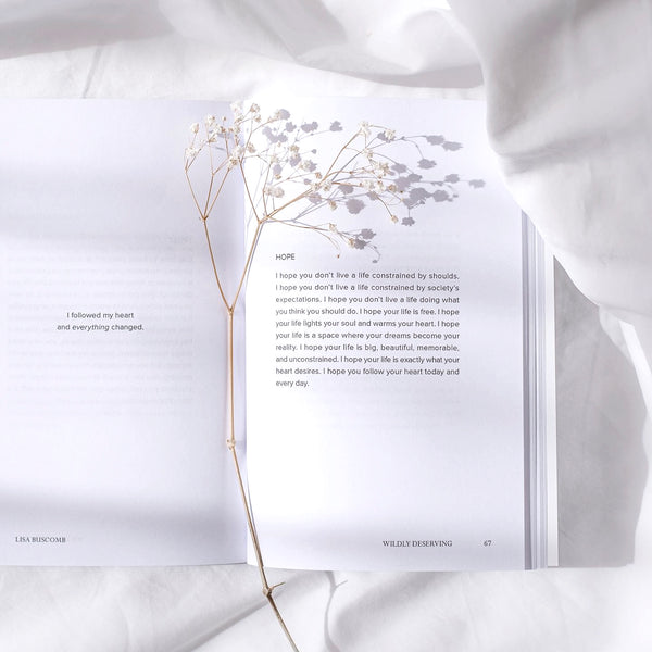 An EVERYDAY MOMENTS Book open on a white bed with a plant on it, showcasing everyday moments of loving yourself and the magic within. (Brand Name: Lisa Buscomb)