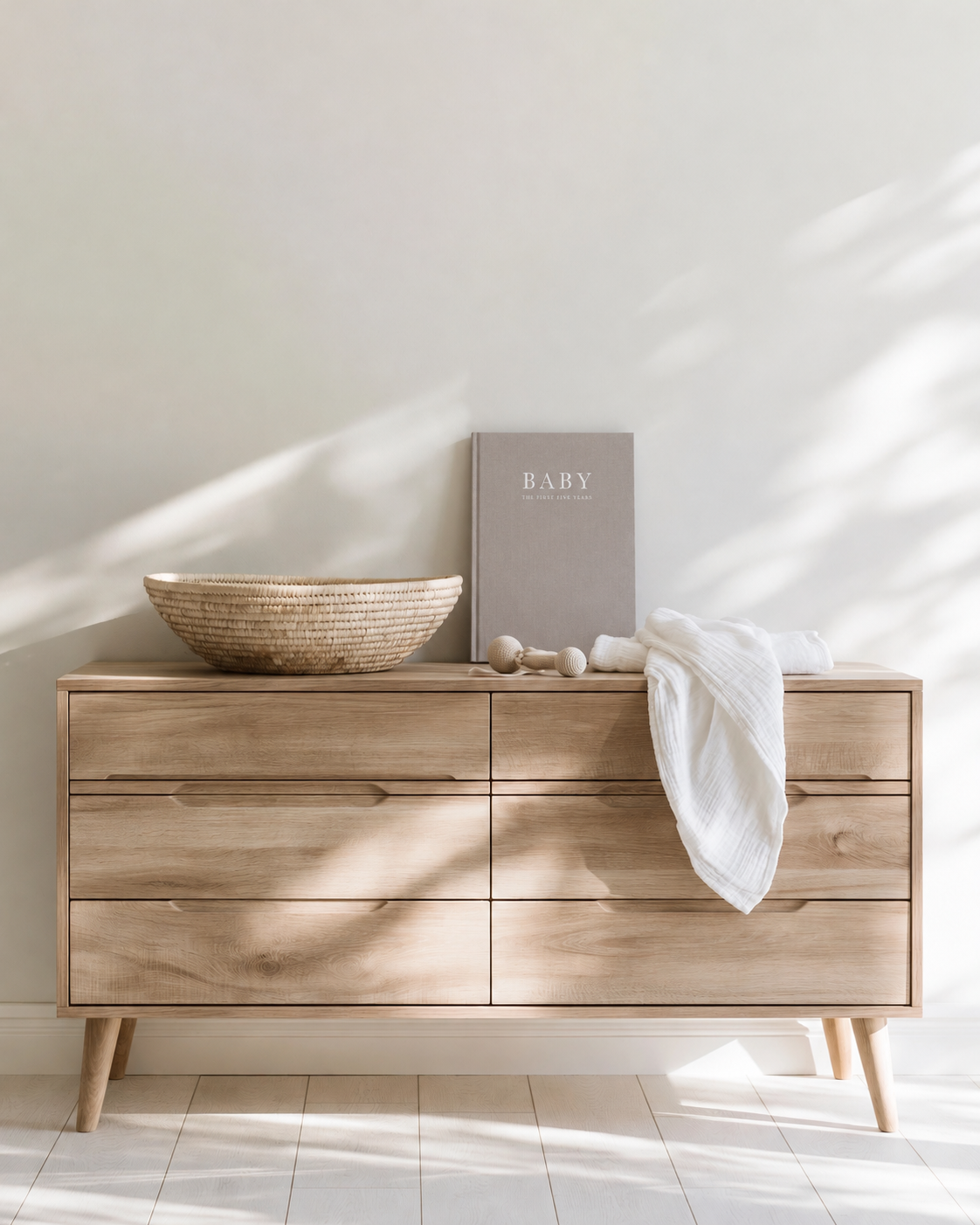 Write To Me Baby memory journal in grey linen standing on an oak nursery dresser with a woven bowl and muslin